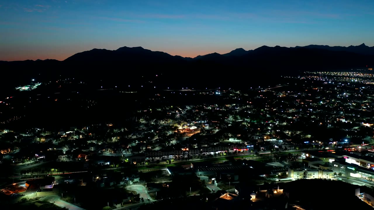 Wide nighttime aerial over Los Cabos, Mexico, with bright road lines, low building clusters, distant lights, and a dark mountain ridge, no visible people.