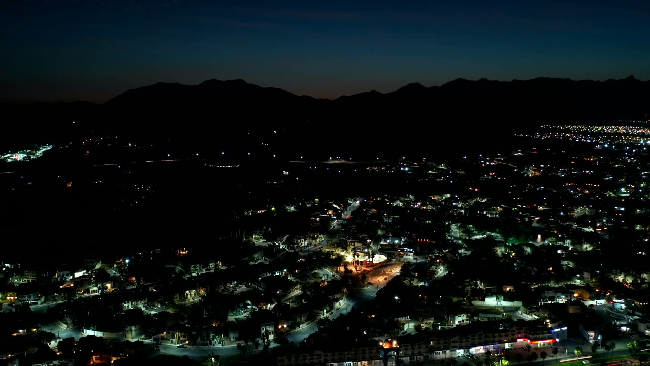 Night aerial frame over Los Cabos, Mexico, showing dense green-white street lighting, a darker central hillside area, and no visible people.