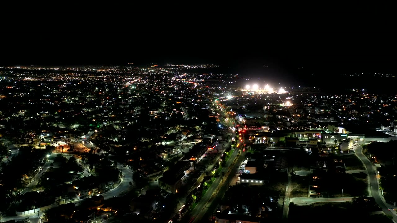 High nighttime view across Los Cabos, Mexico, with a diagonal illuminated road, dense small city lights, dark foreground blocks, and no visible people.