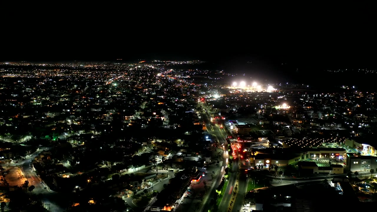 Aerial night view over Los Cabos, Mexico, with scattered city lights, a bright road line, dark sky, and no visible people.
