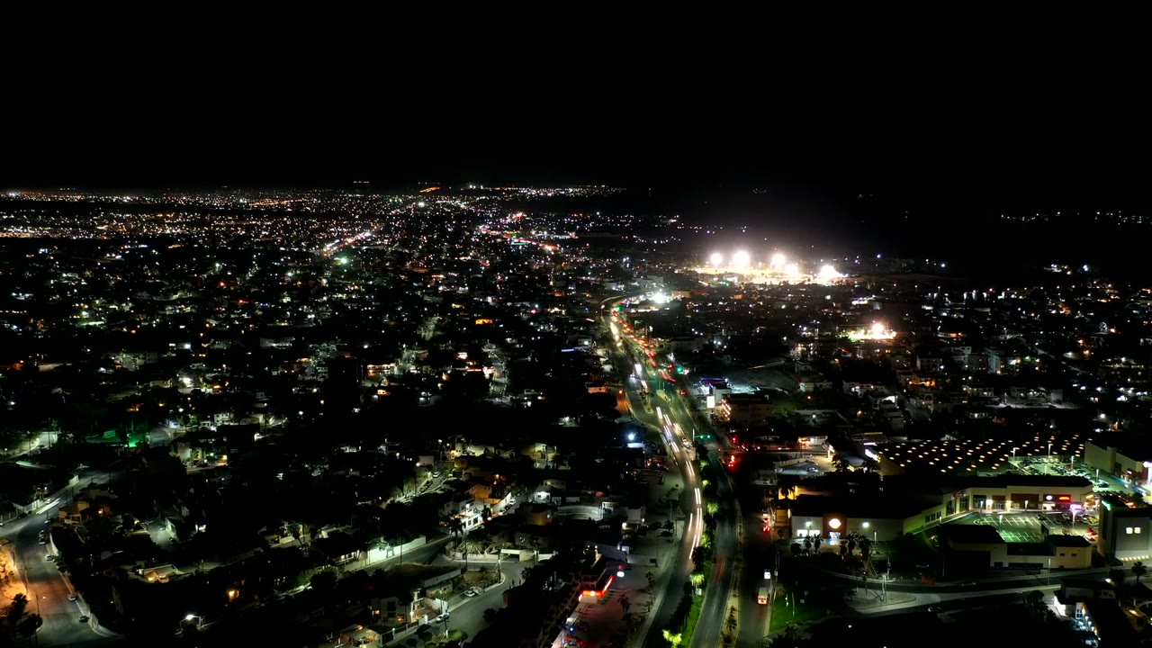 Los Cabos night aerial with a central illuminated road, clusters of buildings, distant bright lights, and no visible people.
