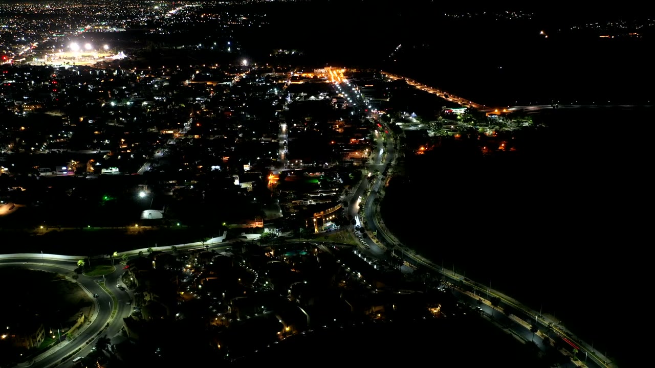 Opening frame of Los Cabos, Mexico at night, with bright road traffic, scattered building lights, dark water to the right, and no visible people.