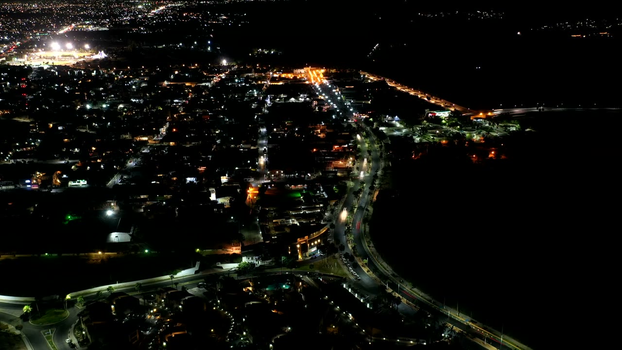Aerial view over Los Cabos, Mexico with bright traffic lines, dense street lighting, dark open water on the right side, and no visible people.