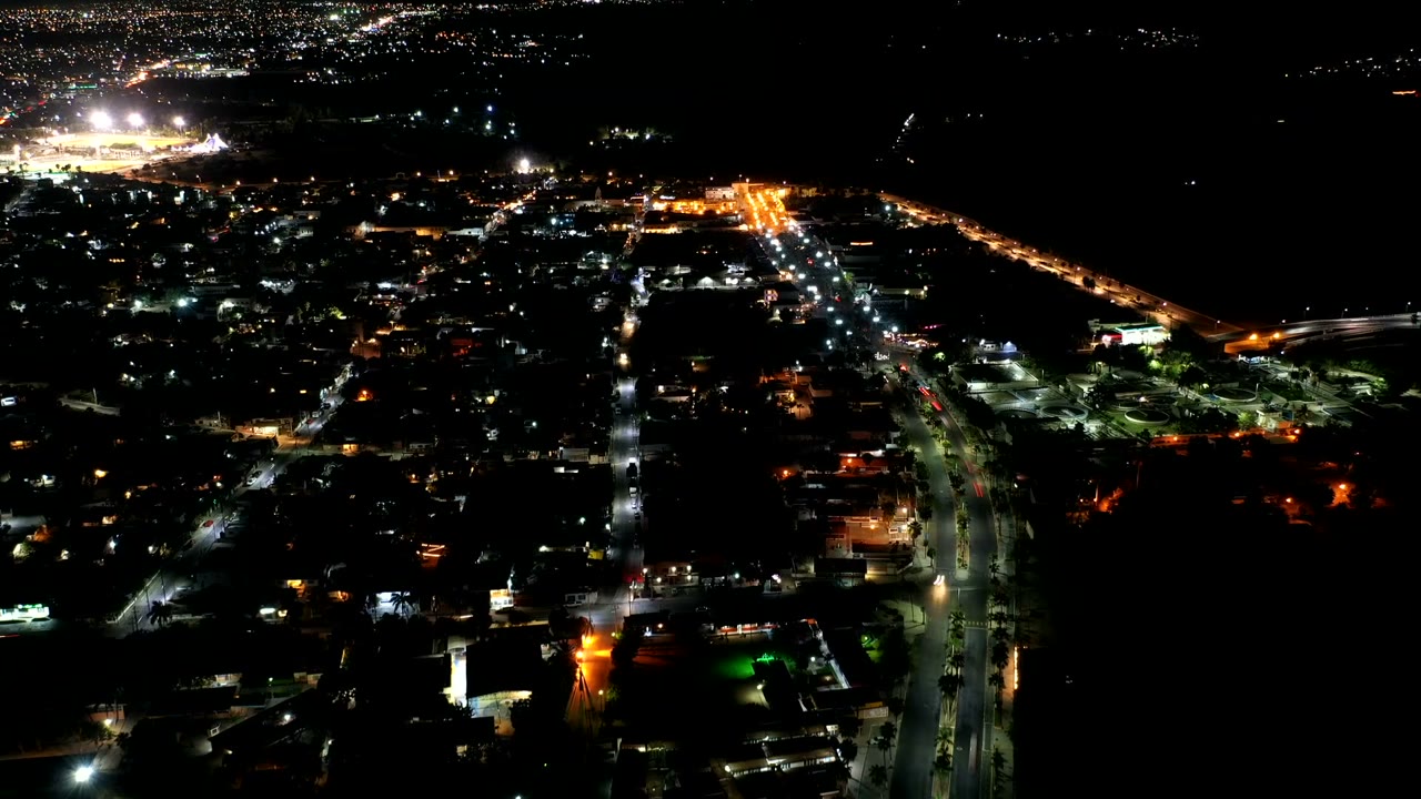 Second frame of the Los Cabos night aerial, with a central vertical road of white lights, orange-lit streets to the right, dark sky, and no visible people.