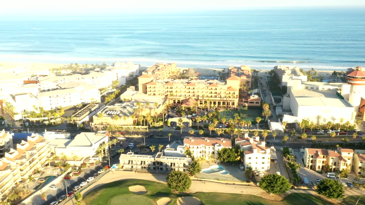 Opening aerial frame over Los Cabos, Mexico, with blue ocean beyond beachfront buildings, palm-lined streets, a golf green in the foreground, and no visible people.