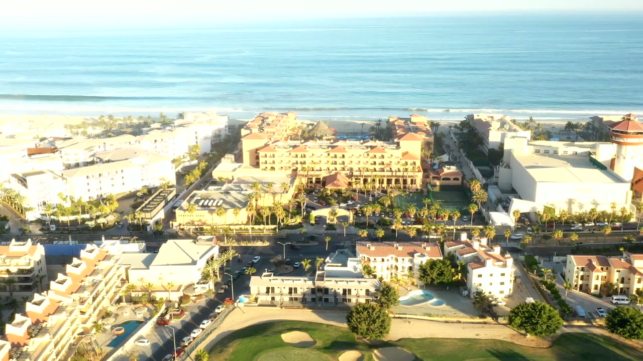 Second aerial frame in Los Cabos, Mexico, showing the ocean horizon, pale surf, white beachfront buildings, terracotta roofs, road traffic, and no visible people.