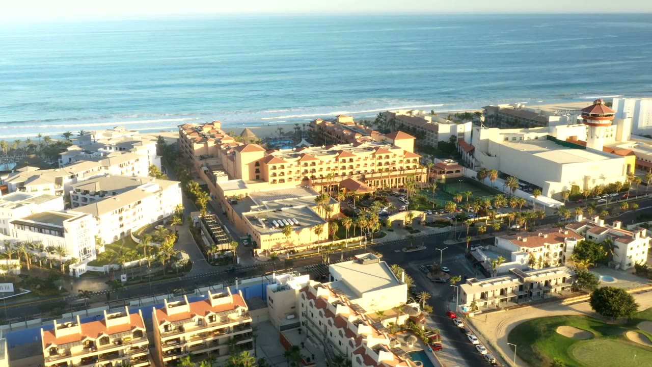 Opening aerial frame over Los Cabos, Mexico with hotels, palm-lined streets, a beach edge, blue ocean, and no visible people.