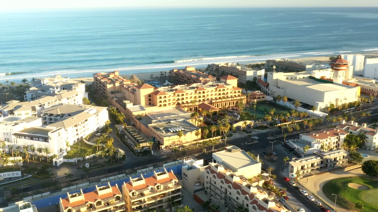 Second aerial frame in Los Cabos, Mexico showing sunlit coastal buildings, cars on nearby roads, layered wave lines, and no visible people.