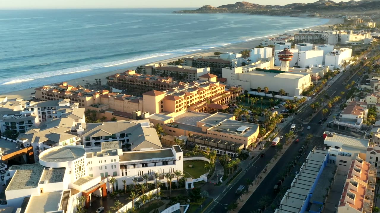 Second aerial frame in Los Cabos, Mexico, showing the beach and blue water to the left, white and terracotta buildings in the center, a straight roadway to the right, and no visible people.