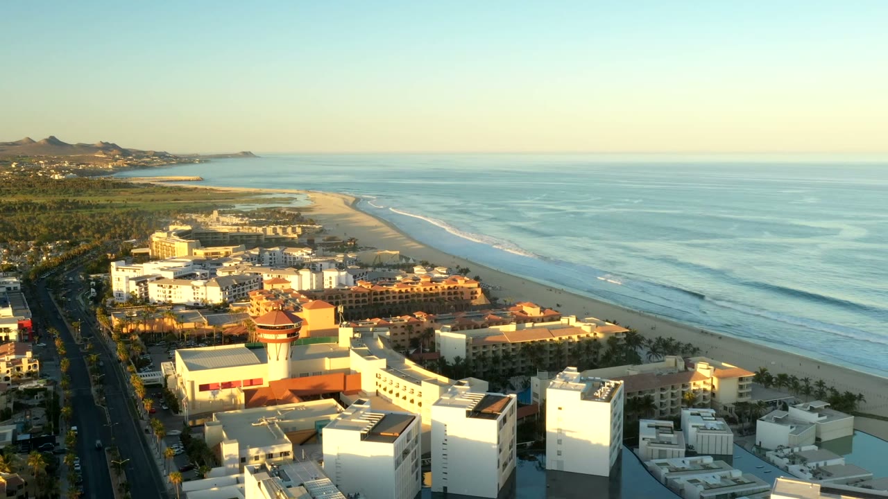 Aerial view over Los Cabos, Mexico with beachfront buildings in the foreground, a long sandy shoreline, blue ocean to the right, distant hills, and no visible people.