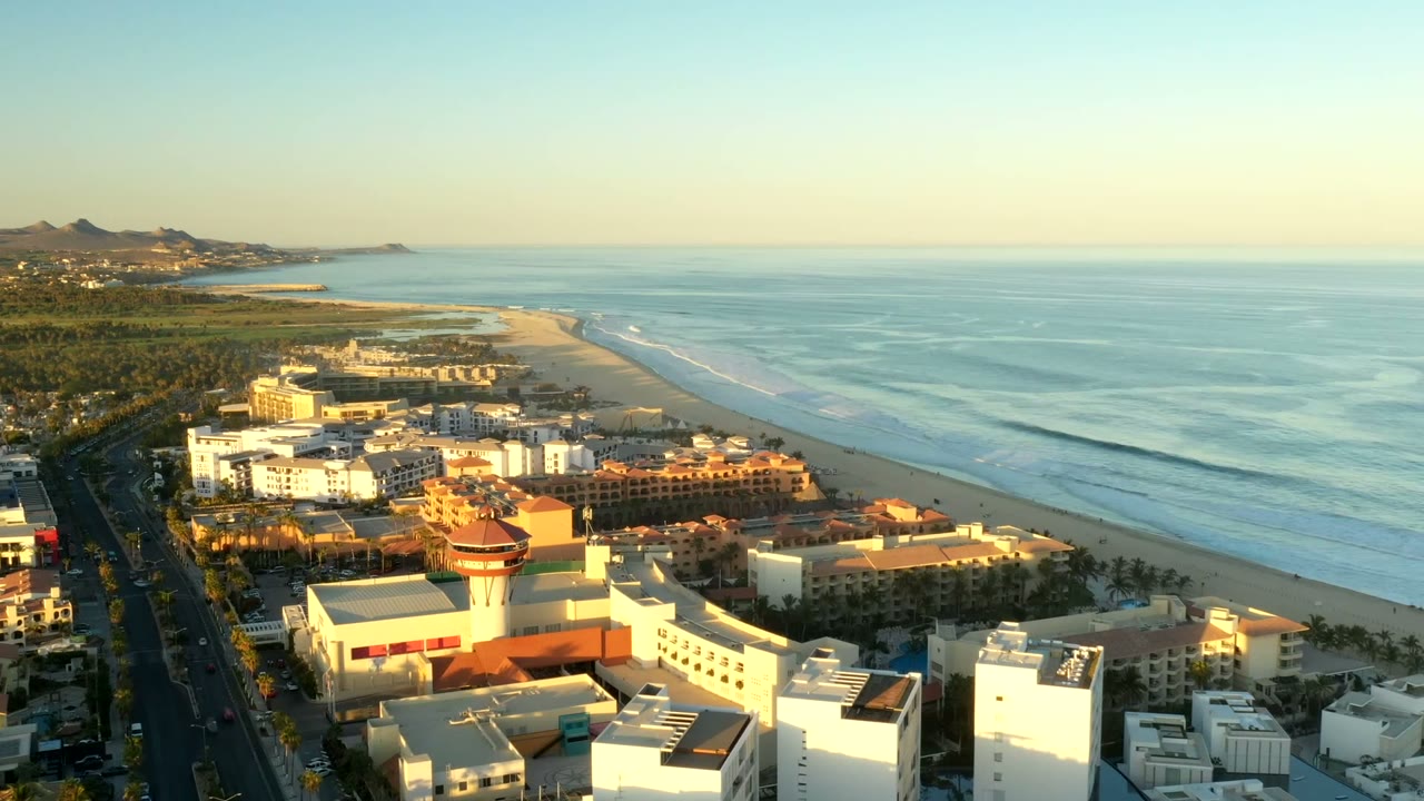 High aerial frame in Los Cabos, Mexico showing a foreground hotel district, a straight road at left, a broad beach, repeating wave bands, hazy horizon, and no visible people.