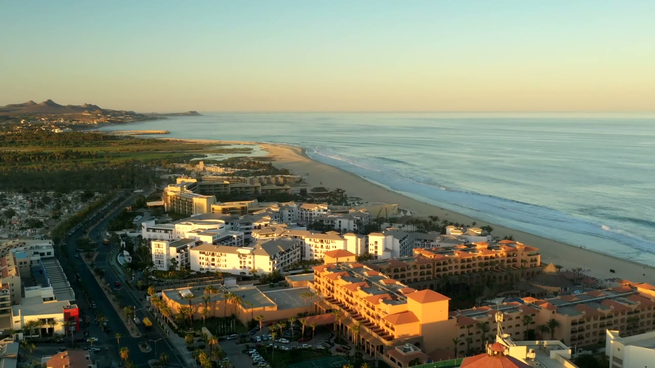 Second aerial frame of Los Cabos, Mexico with foreground hotel blocks, a long beach, parallel surf lines, still blue water, distant hills, and no visible people.