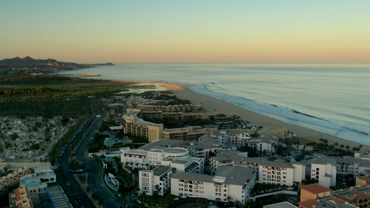 Wide aerial view in Los Cabos, Mexico showing large coastal buildings, a road, beach, ocean surf lines, distant mountains, and no visible people at the start of the clip.