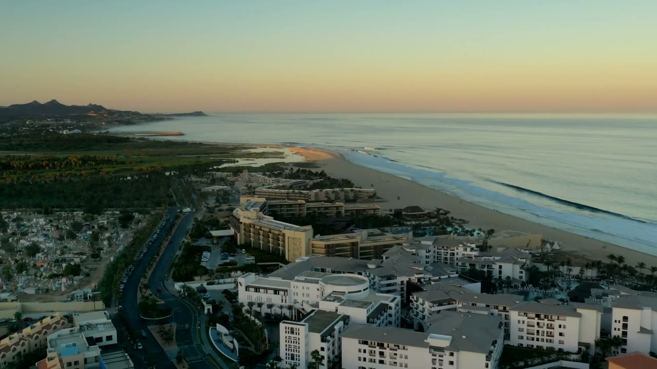 Aerial frame over Los Cabos, Mexico with foreground white buildings and roads, midground shoreline development, broad beach, ocean bands, and no visible people.