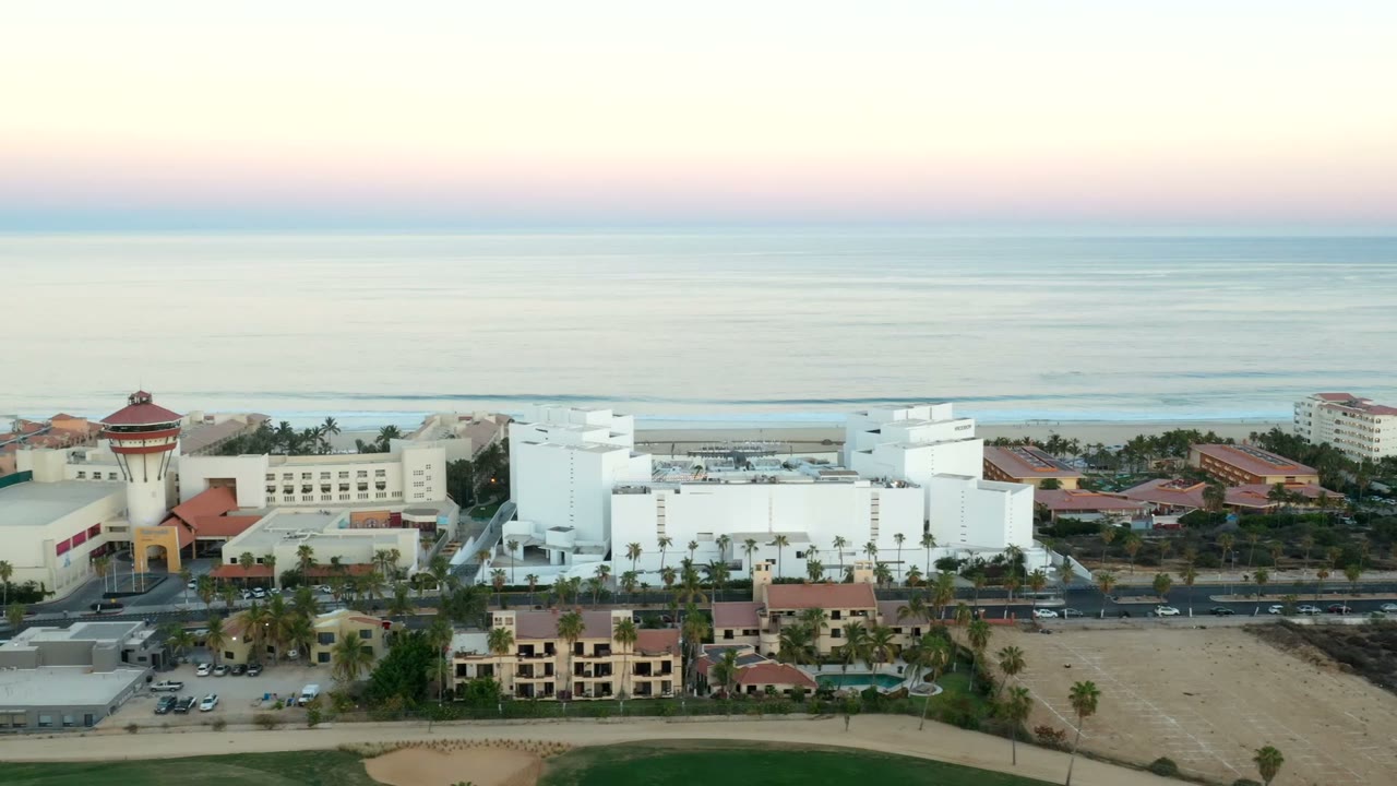 Aerial sunset view over Los Cabos, Mexico with white beachfront buildings, red-tile roofs, roads, parked cars, palms, sandy lots, ocean horizon, and no visible people.
