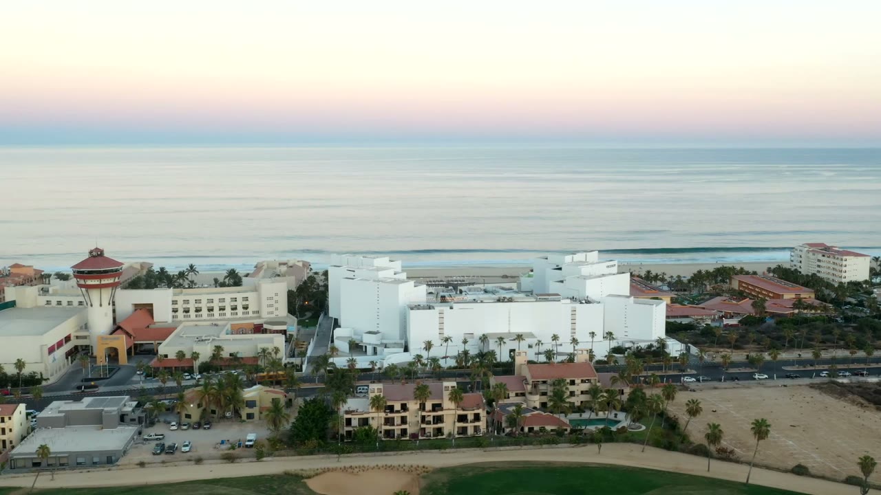 Los Cabos aerial frame with the white building complex near center, a tower-like red-roofed structure, palm-lined roads, cars, beach sand, ocean, and no visible people.