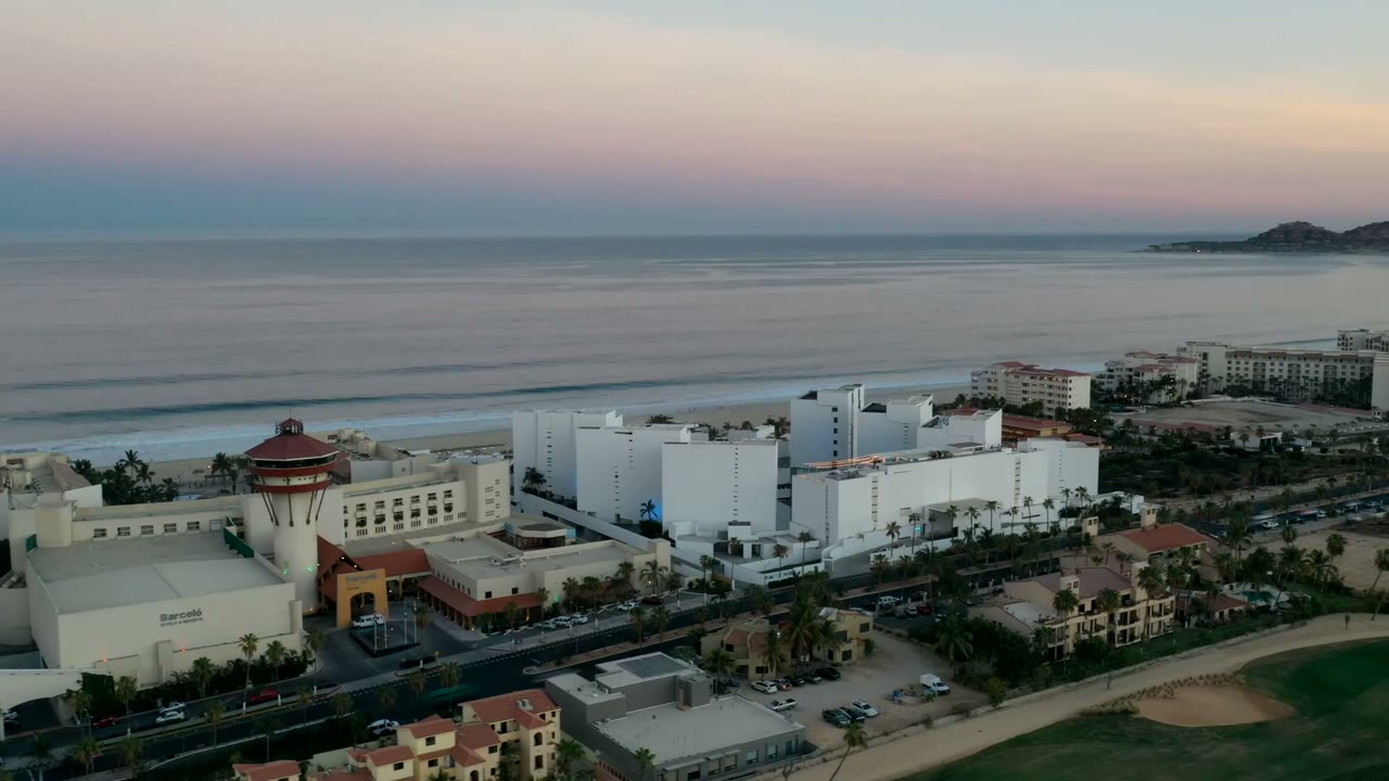 High aerial frame in Los Cabos showing large white buildings, palm-lined streets, sandy beach, calm ocean, distant hills, and no visible people.