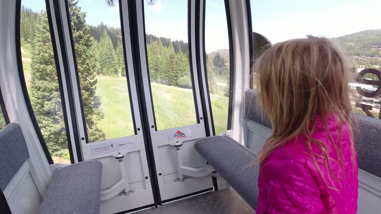At 0:01 in Winter Park, Colorado, the child stands near gray bench seating while tall black window frames divide the view of green hillside and trees outside.