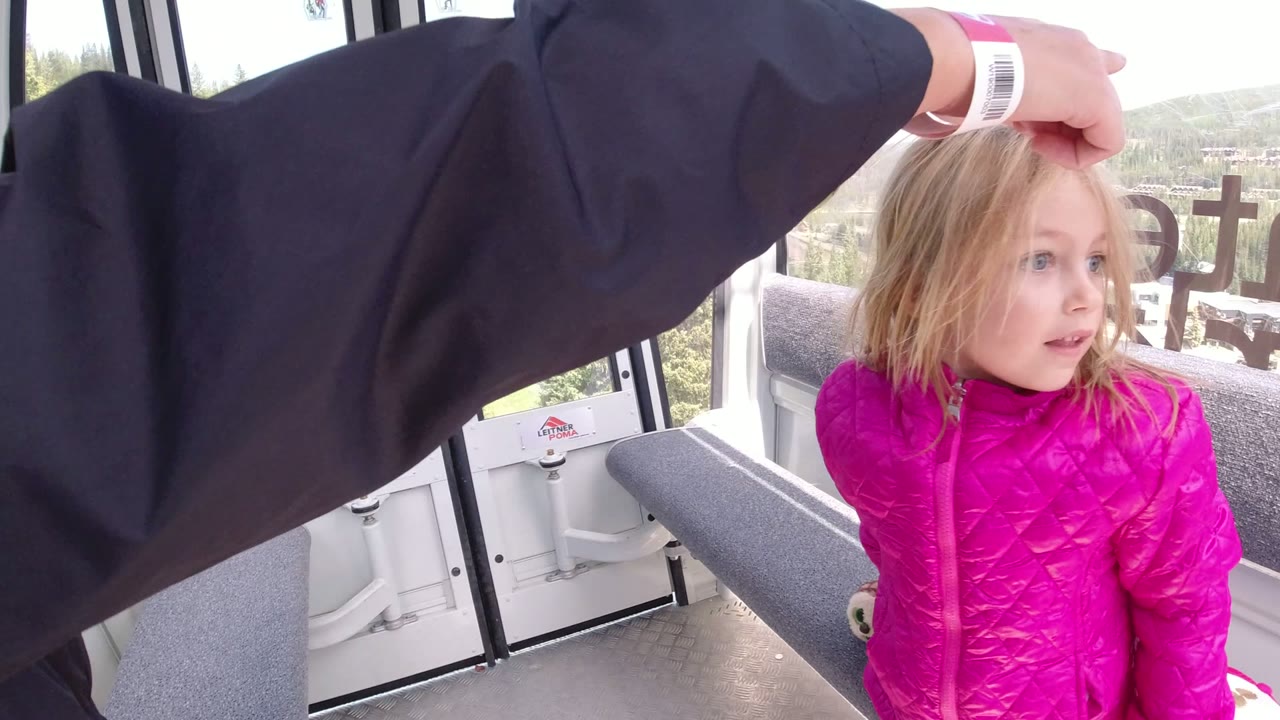 Young girl in a bright pink coat looks across the enclosed ski lift cabin in Winter Park, Colorado, while an adult arm points toward the window.