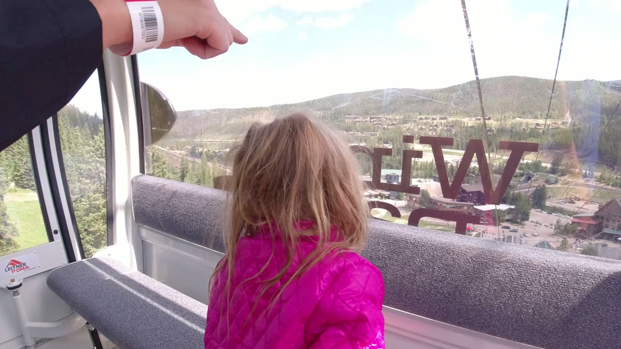 Girl in a pink coat turns toward the Winter Park, Colorado view through the lift window, with reversed lettering, hills, trees, and buildings outside.