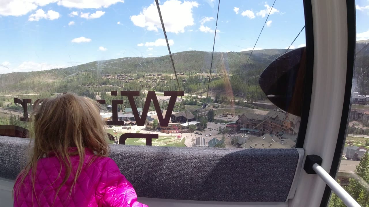 Child in a pink jacket looking out a gondola window over mountain buildings.