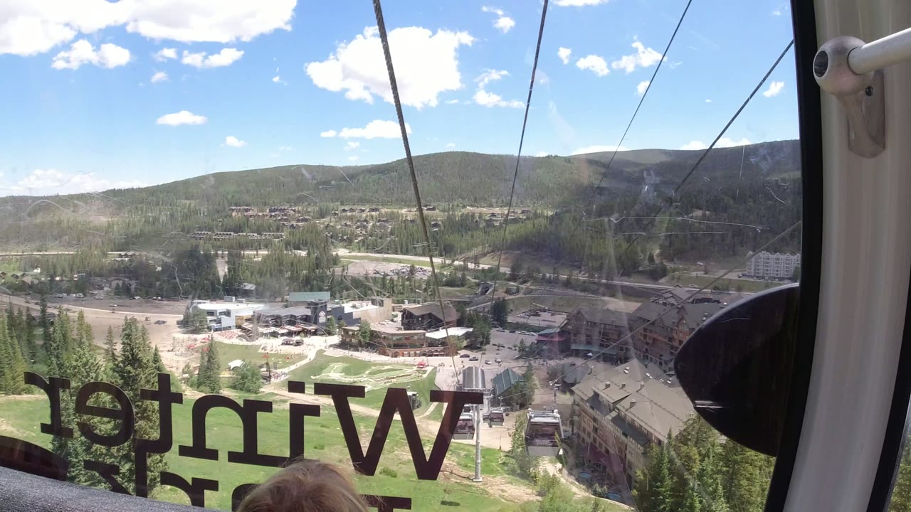 The camera looks through the gondola glass at Winter Park, Colorado, with a small portion of a child's head at the bottom edge, reversed Winter lettering, lift cables, buildings, pine trees, and distant hills.