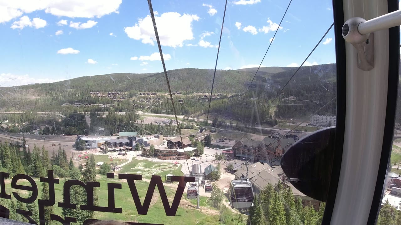 Interior gondola view over Winter Park, Colorado with reversed window lettering, cable lines, resort buildings, evergreen hills, and no visible people.