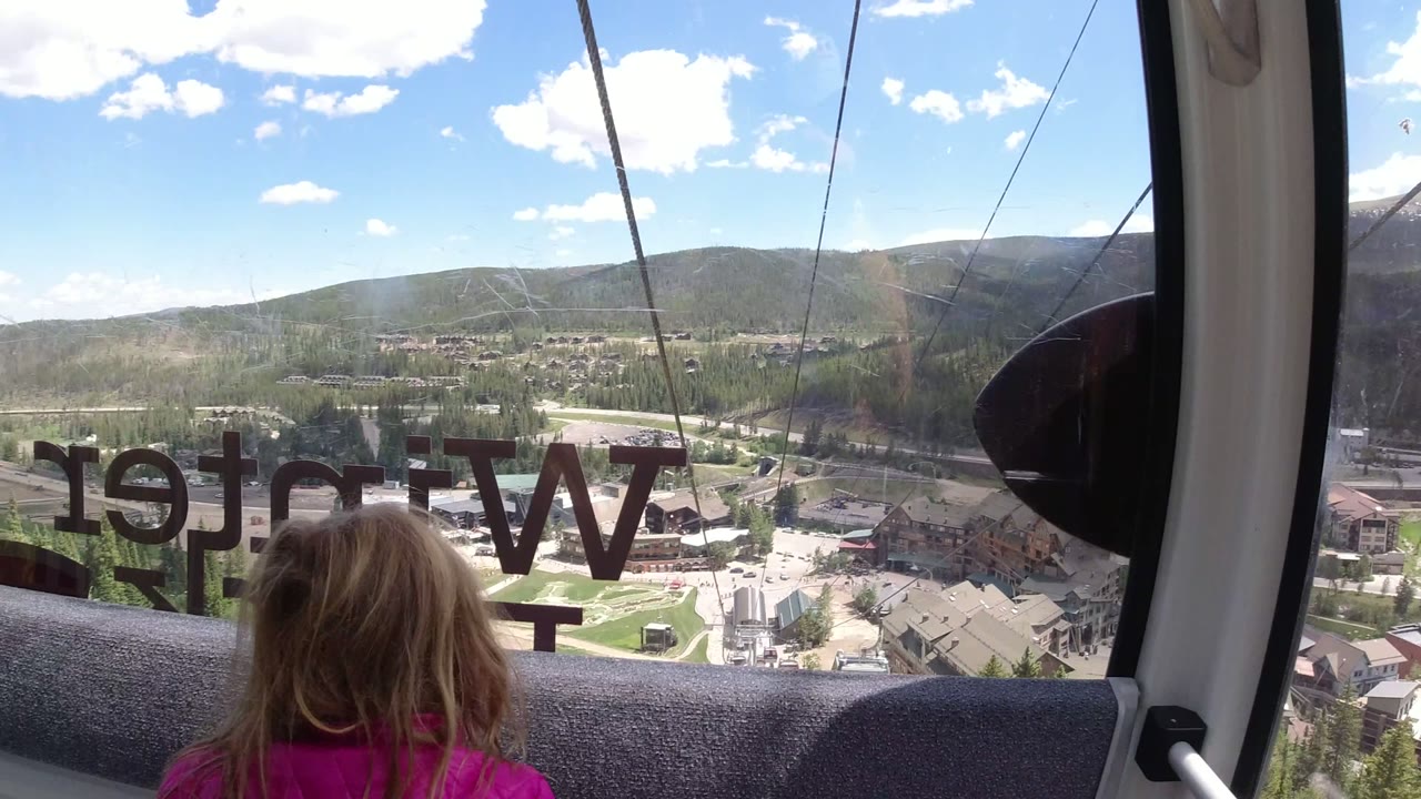 A young child with blond hair sits or stands inside a gondola in Winter Park, Colorado, facing the window with reversed lettering and mountain terrain beyond.