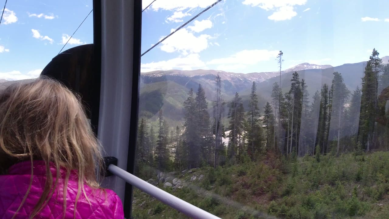 At 0:01 in Winter Park, Colorado, the child remains near the gondola glass while dark window framing, overhead cable lines, green slopes, and distant ridges fill the view.