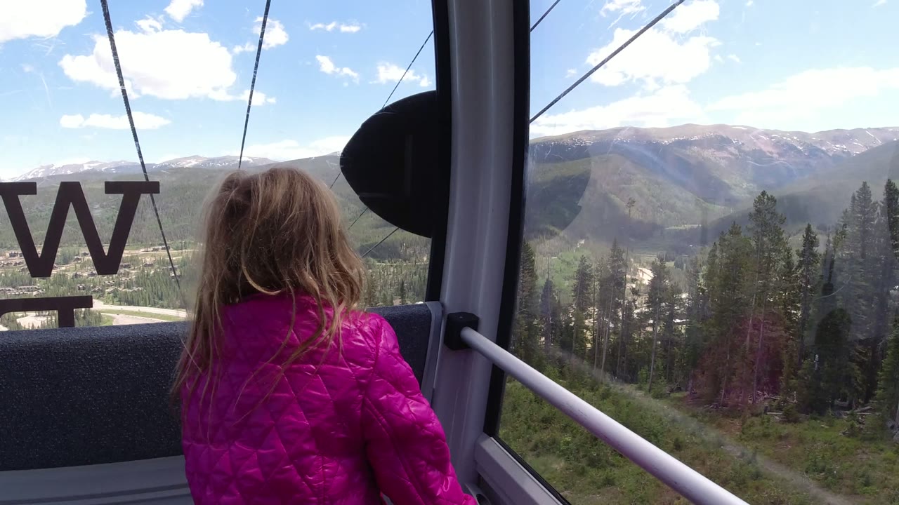 A second of the Winter Park air cab view shows the child centered lower in frame, reversed window letters on the glass, and forested mountains beyond.