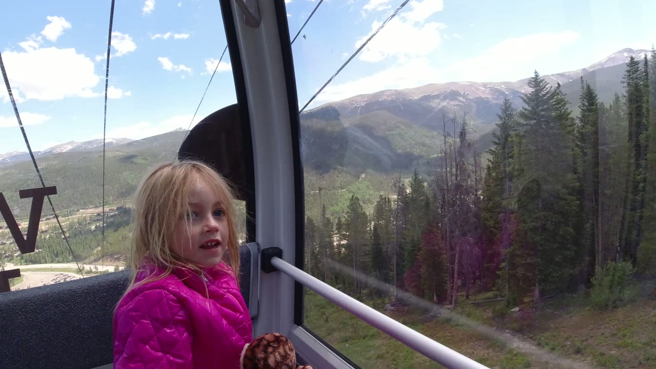 Blonde child in a pink jacket inside a cableway cabin with mountains outside the window.