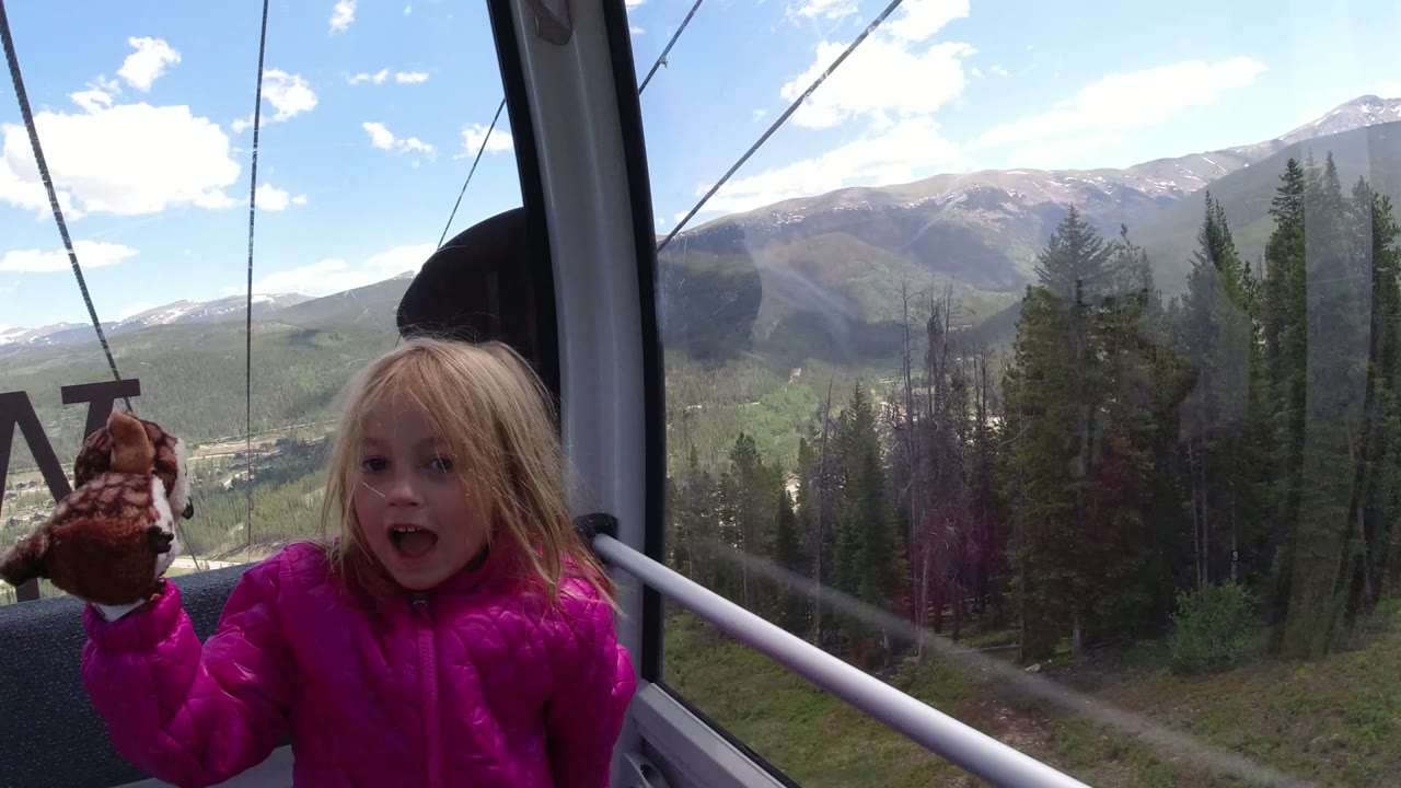 The child raises a small plush toy while standing inside the Winter Park cableway cabin, with the forested slope and mountain ridge seen through the large windows.