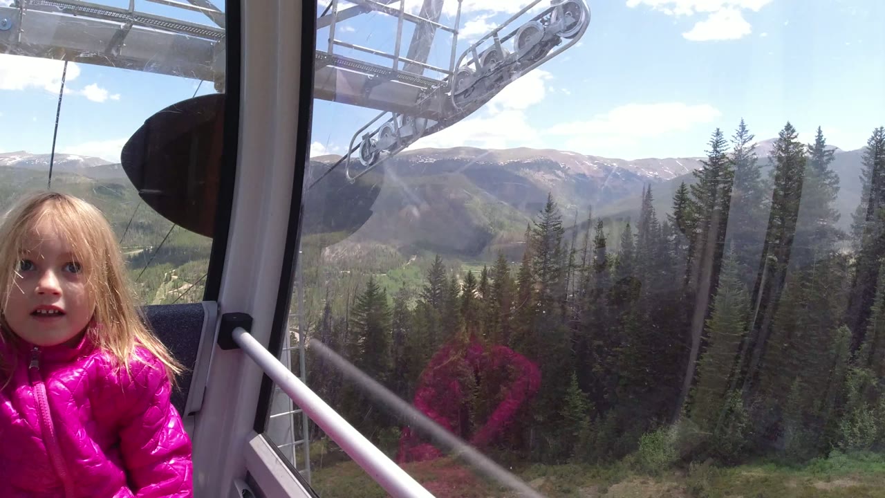 The child remains seated by the gondola window in Winter Park, Colorado, while lift hardware, cable lines, evergreen forest, and mountain ridges fill the view outside.