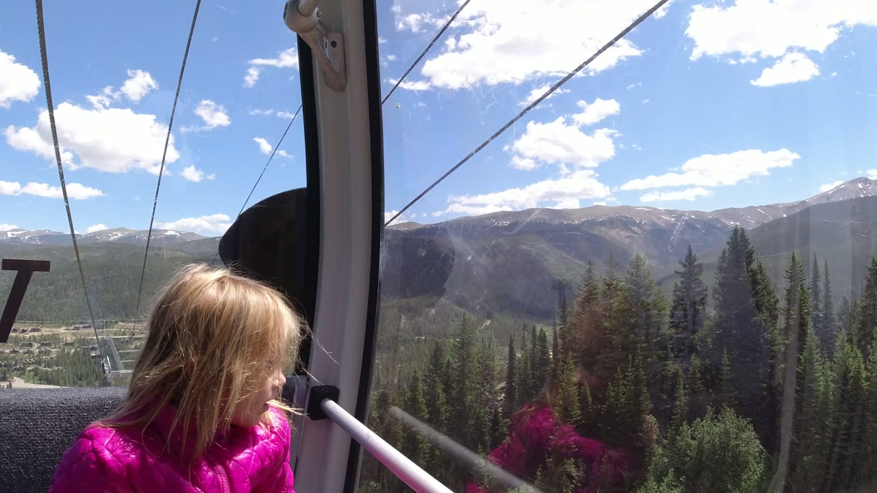 A child in a pink jacket sits near the left side of a cable car window in Winter Park, Colorado, with lift cables, evergreen slopes, distant mountains, and bright clouds outside.