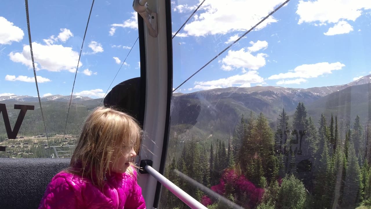 The child remains seated inside the Winter Park cable car while the window frames a forested valley, lift infrastructure, distant ridgelines, and reflected lettering on the glass.