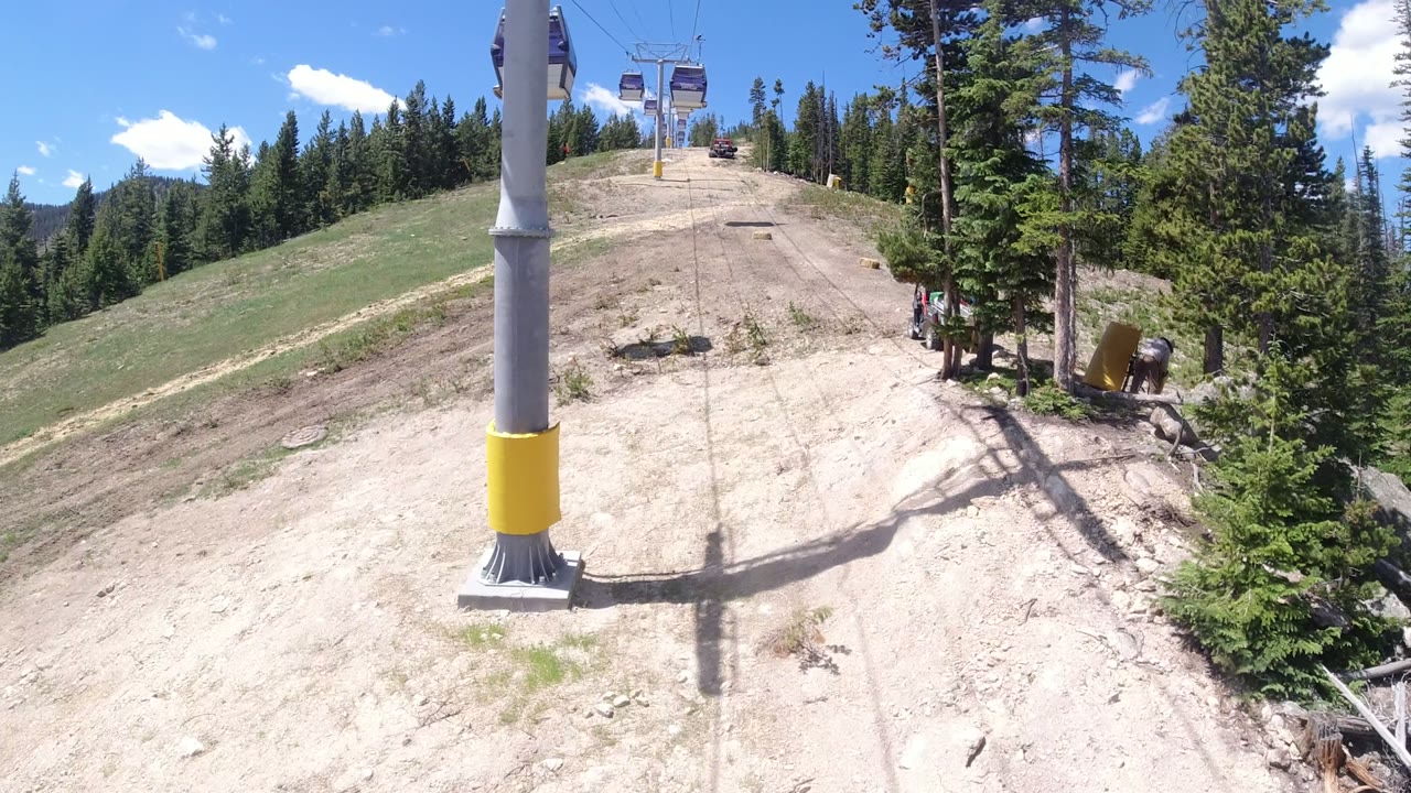 The lift tower sits near center frame in Winter Park, Colorado, with its shadow falling across the dry uphill track, pine trees at right, and no visible people.
