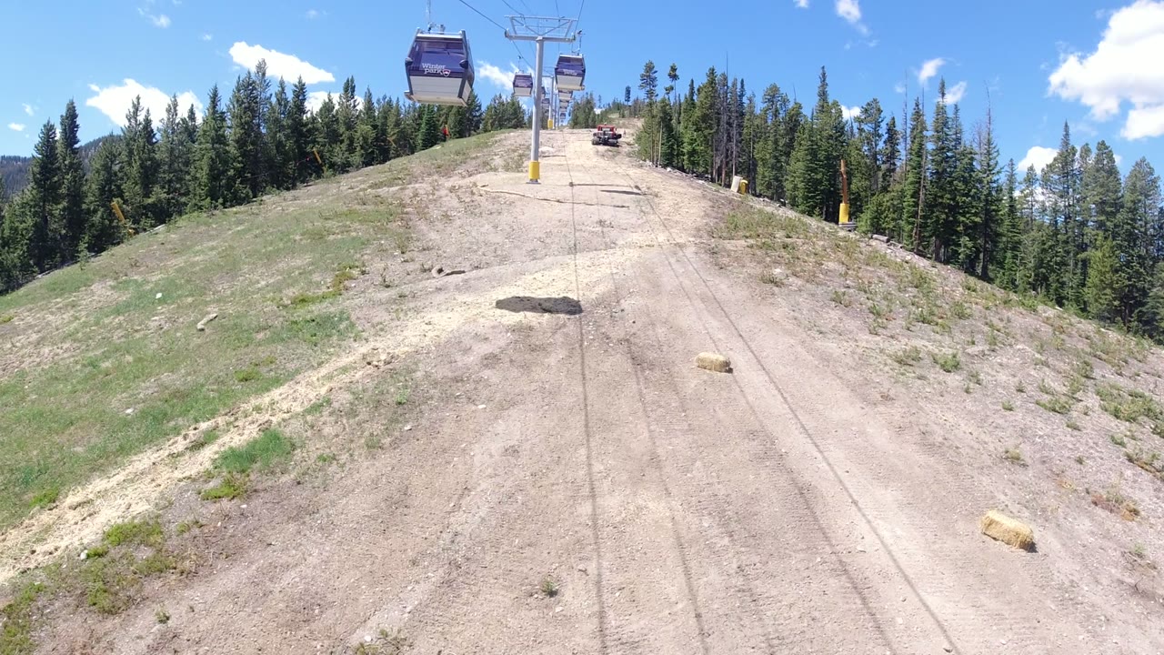 The Winter Park lift line continues uphill with a metal tower near the center, dirt service tracks on the slope, pine trees framing the scene, and no visible people.