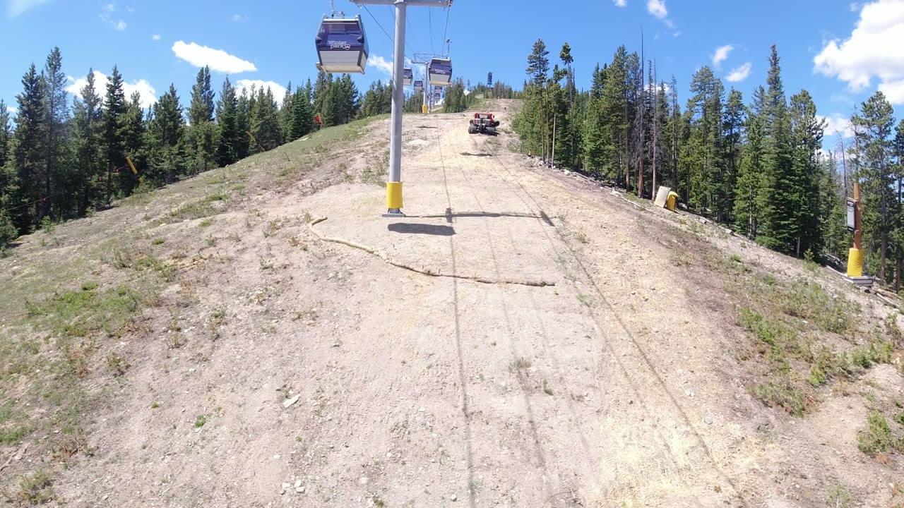 A Winter Park, Colorado lift line continues uphill over a dry graded run, with gondola cabins near the top of frame, forest edges, scattered clouds, and no visible people.