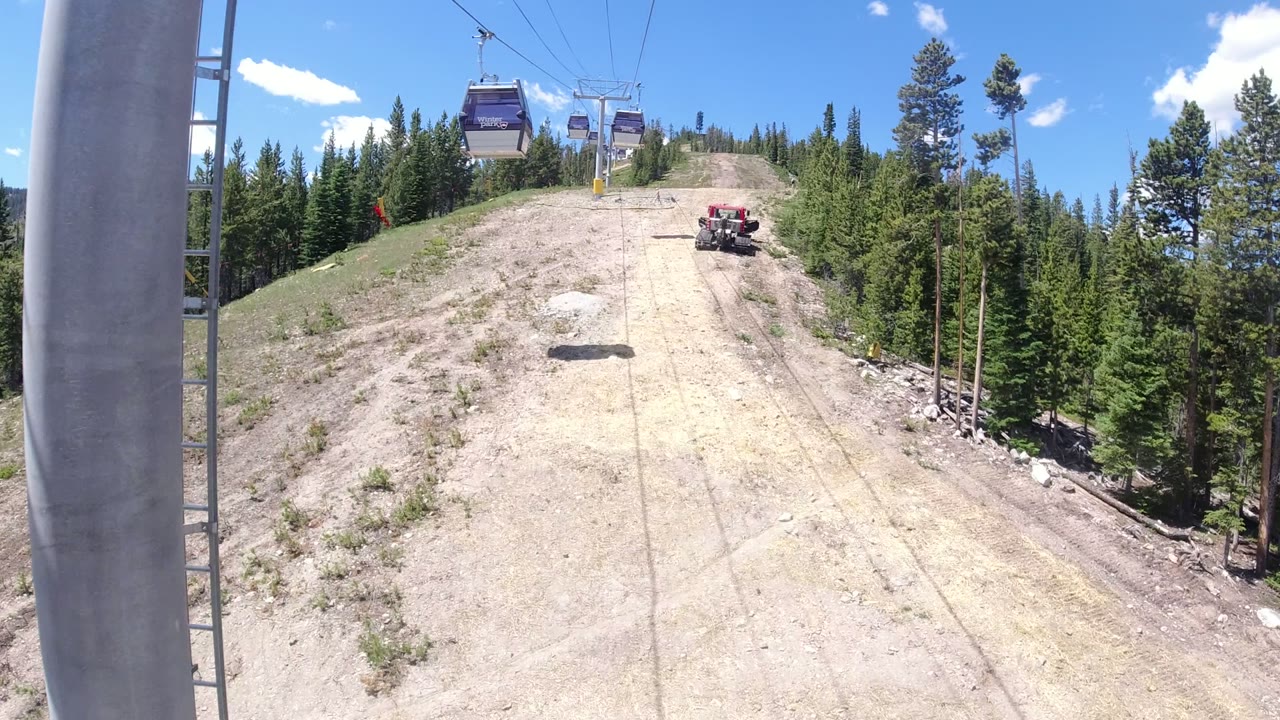 Winter Park, Colorado slope seen from an elevated lift-line angle, with gondola cabins overhead, a red utility vehicle on the run, and evergreen trees along the right side.