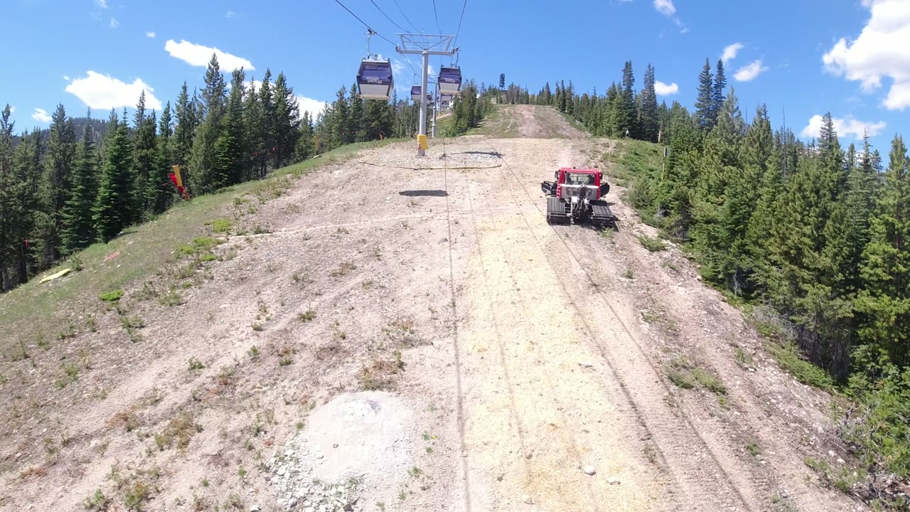 A ropeway cabin hangs near the top of frame over a cleared slope in Winter Park, Colorado, with evergreen forest on both sides and a red tracked tractor parked on the right.