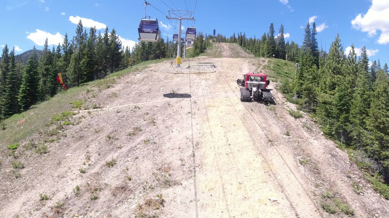 The ropeway cabin sits high over the Winter Park, Colorado slope while the red tracked tractor remains on the right edge and dense evergreens frame the cleared run.
