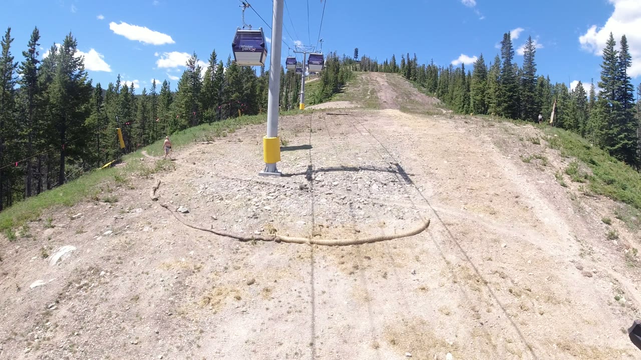 A gray lift tower with yellow padding stands beside the cable line in Winter Park, Colorado, with gondola cabins overhead, forested slopes beyond, and a small distant person near the left edge.