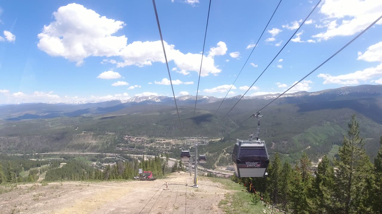 Multiple gondola cabins continue along overhead cables in Winter Park, Colorado, with a lift tower, cleared slope, forested valley, distant mountains, bright clouds, and no visible people.