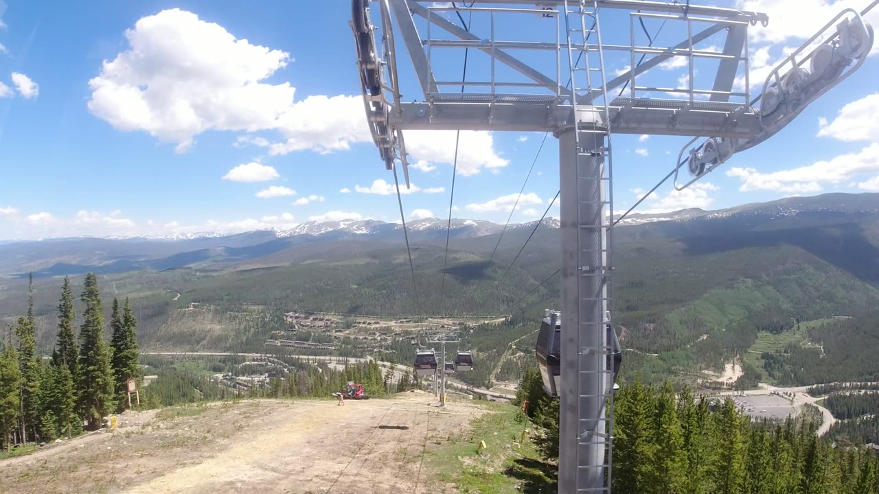 A steel ropeway tower and overhead cable assembly fill the upper frame above a dirt slope, evergreens, and distant mountains in Winter Park, Colorado, with no visible people.