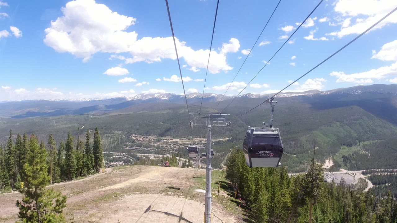 A gondola cabin hangs farther down the cable line over conifer forest and open slope terrain, with Winter Park, Colorado mountain ridges and bright clouds in the background, no visible people.