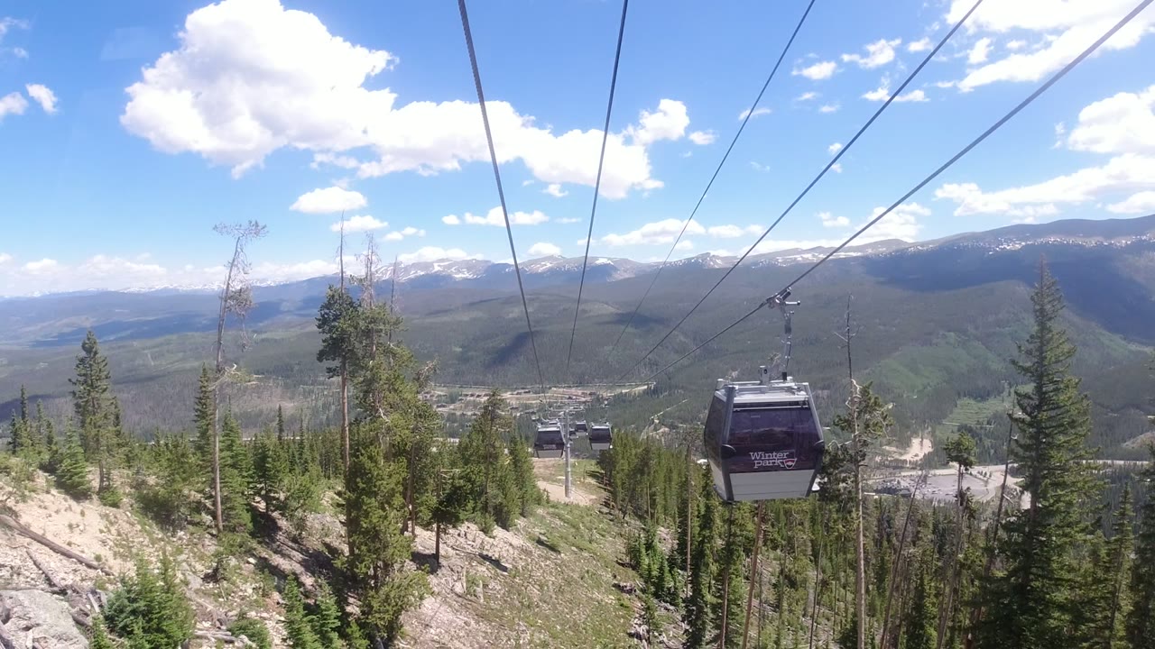 A dark Winter Park gondola cabin passes close to the camera on overhead cables, with forested slopes and layered mountains behind it, and no visible people.