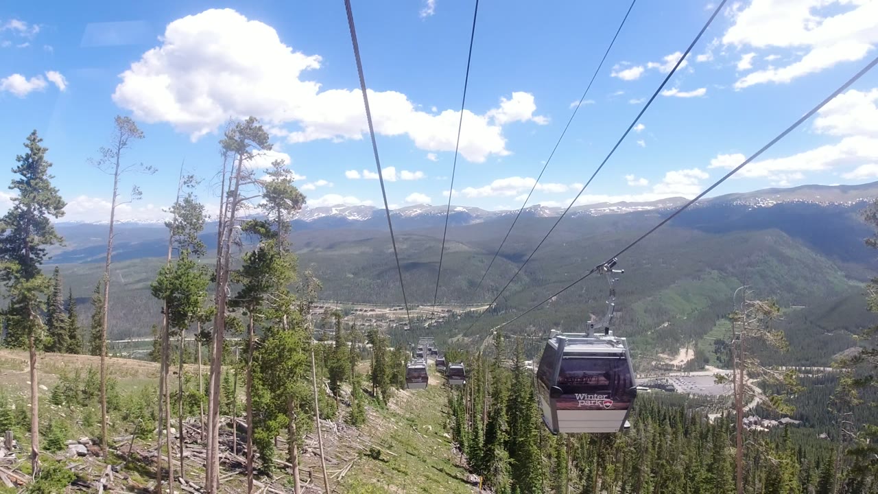 A gondola cabin marked for Winter Park moves near the camera over trees and a distant mountain valley, with no visible people.