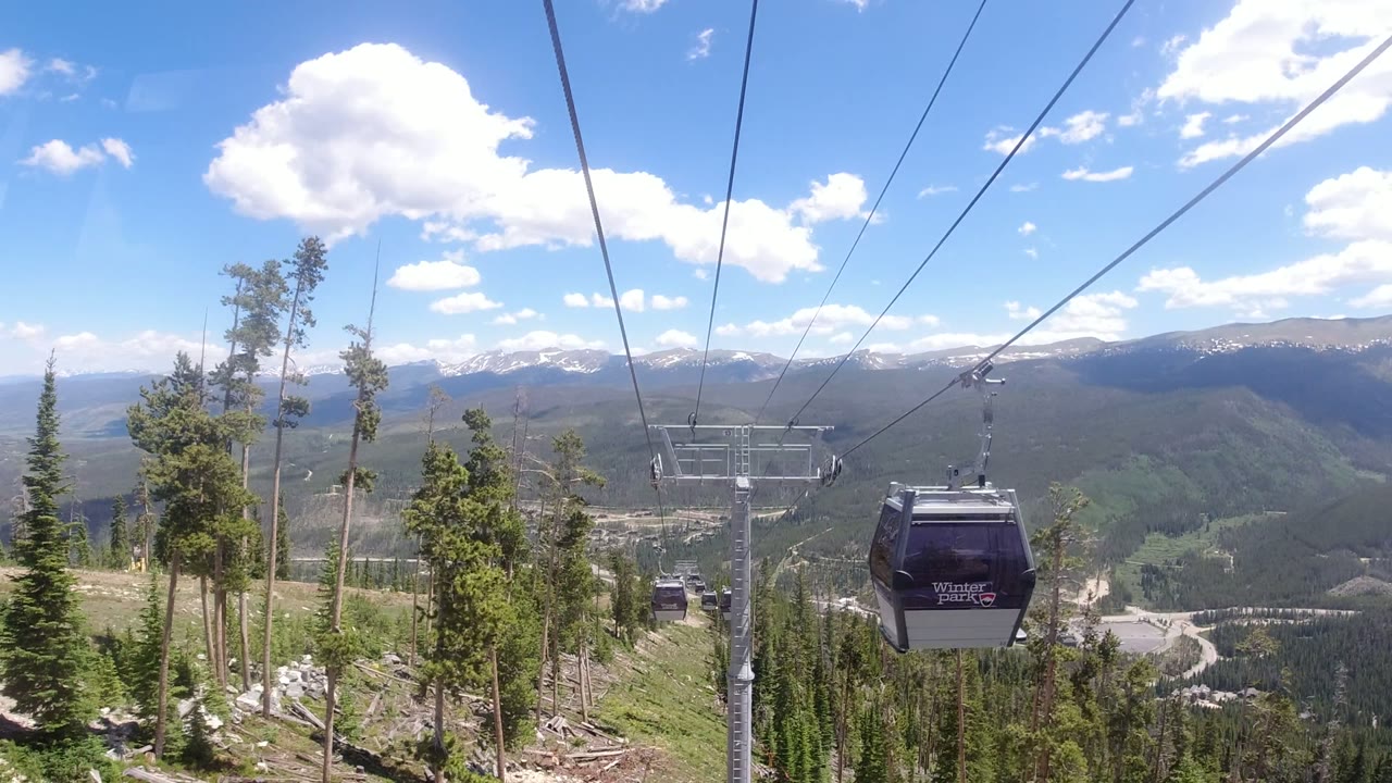 A black gondola cabin passes close in the foreground in Winter Park, Colorado, with cables, a lift tower, pine forest, and distant mountains behind it, with no visible people.