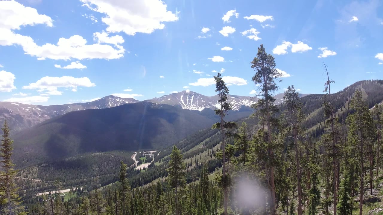 Winter Park, Colorado slopes and a pale valley road appear between foreground evergreens, with distant mountains and no visible people.