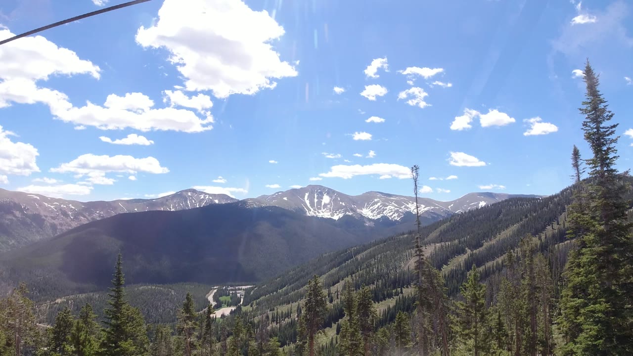Wide mountain valley with evergreen trees, distant snow patches, blue sky, and cable line overhead.